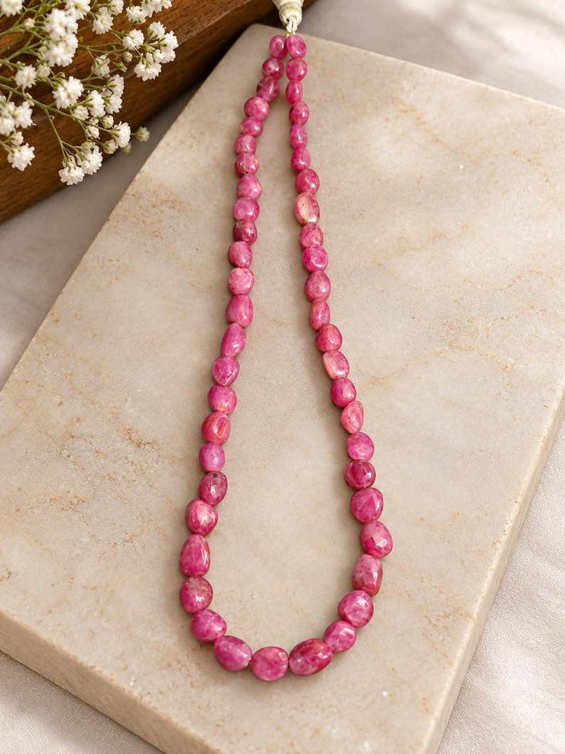 May include: A single strand necklace composed of numerous small, faceted, irregular pink gemstone beads. The beads are strung on a light-coloured cord, creating a continuous loop of vibrant, polished stones with varying shapes and sizes.