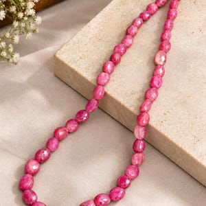 May include: A necklace composed of numerous faceted, oval-shaped pink gemstone beads strung together. The beads exhibit a vibrant magenta hue with natural variations in texture and colour, creating a continuous strand that loops gracefully.