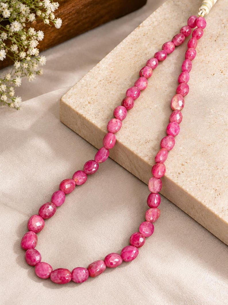 May include: A necklace composed of numerous faceted, oval-shaped pink gemstone beads strung together. The beads exhibit a vibrant magenta hue with natural variations in texture and colour, creating a continuous strand that loops gracefully.