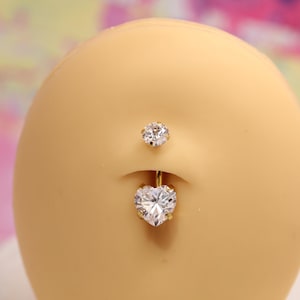 Könnte beinhalten: Ein goldfarbener Bauchnabelring mit einem herzförmigen, klaren Kristallanhänger. Ein kleinerer, runder, klarer Kristall befindet sich oberhalb des Nabels. Der Schmuck wird auf einer hellgelben Oberfläche präsentiert.