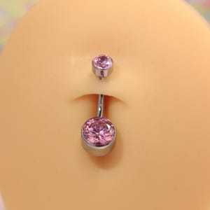 Può includere: Un piercing all'ombelico con una grande gemma rosa rotonda nella parte inferiore e una gemma più piccola abbinata nella parte superiore. Il gioiello è color argento e posto su un incarnato chiaro.