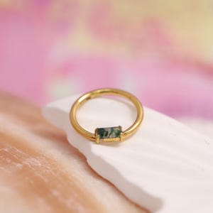 Peut inclure: Une bague dorée ornée d'une pierre verte rectangulaire. La bague repose sur une surface blanche, sur un fond rose et jaune doux. La pierre est de forme rectangulaire et est placée au centre de la bague.