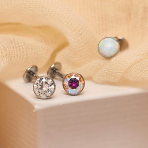 May include: Three metal labret studs with flat back posts. One stud features a clear faceted stone, another has a multi-colored iridescent stone with a deep pink center, and the third displays a smooth, round, milky white opal-like cabochon.