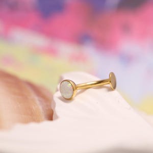 May include: A gold-colored labret stud with a round, white opal stone. The jewelry piece has a flat back and a curved post. The opal has a pearly, iridescent appearance. The background is a soft, colorful gradient.