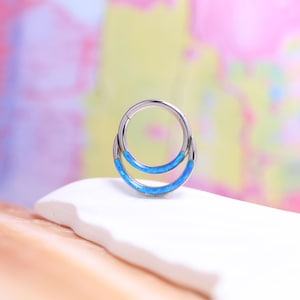 Op de afbeelding: Een zilverkleurige neusring met dubbele hoepel en een blauwe opaal inleg. De sieraden zijn geplaatst op een wit oppervlak met een kleurrijke, vage achtergrond. De hoepel is ontworpen als septumring.