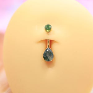May include: A gold-colored belly button ring with a green gemstone at the top and a teardrop-shaped green gemstone pendant. The jewelry is displayed on a light-colored surface.