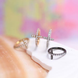 May include: Four rings with cross designs. One is gold-toned, one is silver-toned, and one is black-toned. All three have small, clear stones. The fourth ring is gold-toned with a cross design and small, clear stones. The rings are displayed on a white surface.