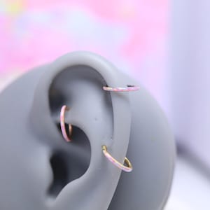 May include: Three gold-colored hoop earrings with pink and white opal-like inlays are displayed on a gray ear model. One earring is in the upper ear, and two are in the lower ear.