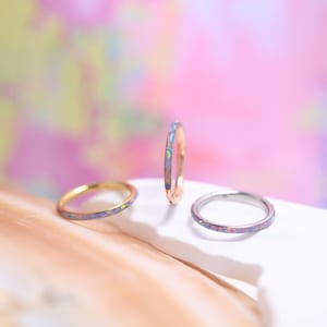 Pode incluir: São apresentados três anéis com incrustações semelhantes a opala. Um anel é dourado, um é de ouro rosa e outro é prateado. Os anéis são colocados sobre um fundo macio e colorido, destacando suas qualidades iridescentes. Os anéis são delicados e elegantes.