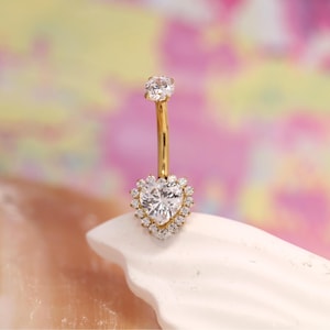 May include: A gold-colored belly button ring with a heart-shaped, clear gemstone pendant. The pendant is surrounded by smaller, clear gemstones. The top of the ring features a single, clear gemstone. The jewelry is displayed on a white surface against a colorful background.
