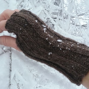 May include: Brown knitted fingerless gloves with a cable knit pattern. The gloves are worn on a hand against a snowy background.