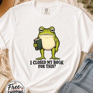 Peut inclure: Un t-shirt blanc à manches courtes avec un motif représentant une grenouille verte tenant un livre vert foncé. Sous la grenouille, le texte indique : "I CLOSED MY BOOK FOR THIS?". Le t-shirt présente un col rond et une coupe classique.