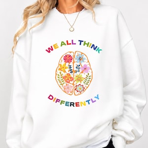 Könnte beinhalten: Ein weißes Sweatshirt mit Rundhalsausschnitt und einer zentralen Grafik eines menschlichen Gehirns mit orangefarbenen Umrissen, das mit bunten Blumen und Blättern gefüllt ist. Der Text WE ALL THINK DIFFERENTLY steht in bunten Buchstaben.
