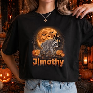 以下が含まれることがあります： 黒いTシャツで、大きなオレンジ色の満月、お化け屋敷のシルエット、枯れ木、3つのカボチャの前を歩くアライグマのグラフィックが描かれています。その下には、オレンジ色の滴るようなフォントで「Jimothy」という名前がプリントされています。