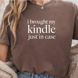 Puede incluir: Una camiseta marrón de manga corta con el texto blanco "i brought my kindle just in case" impreso en una fuente serif en el pecho. La prenda tiene un cuello redondo y una apariencia ligeramente descolorida en toda la tela.
