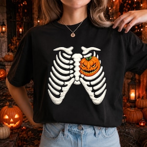 May include: A black t-shirt featuring a white graphic of a human rib cage. Inside the rib cage, on the left side, is a bright orange jack-o'-lantern pumpkin with a carved, menacing face, green stem, and glowing yellow eyes and mouth.