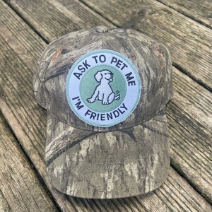 May include: A camouflage baseball cap featuring a circular patch on the front. The patch has a light green background with a white border and a black line drawing of a dog. The text reads ASK TO PET ME I'M FRIENDLY in black capital letters.