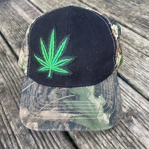 May include: A baseball cap featuring a solid black front panel with an embroidered bright green cannabis leaf patch. The brim and side panels are patterned with a brown, green, and black woodland camouflage print, and the cap has white stitching.