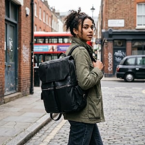 Könnte beinhalten: Ein schwarzer Lederrucksack mit einer großen oberen Klappe, zwei vertikalen Reißverschlusstaschen auf der Vorderseite und verstellbaren Schultergurten. Die Tasche hat eine strukturierte rechteckige Form mit sichtbaren Nähten und silberfarbenen Metallreißverschlüssen.