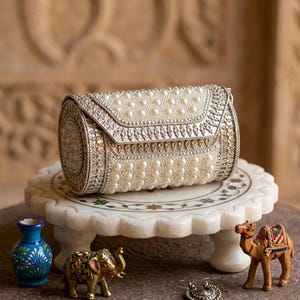 Op de afbeelding: Een zilveren clutch met een parel- en kralenontwerp, rustend op een witte marmeren standaard. De clutch heeft een afgeronde vorm en een decoratieve sluiting. Kleine beeldjes van een olifant en een kameel staan in de buurt.
