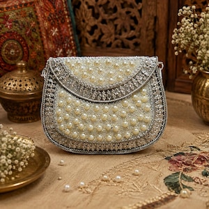 May include: A semi-circular handbag featuring a silver metallic frame and intricate silver embroidery. The exterior is densely decorated with white faux pearls arranged in a repeating pattern across the flap and the main body of the bag.