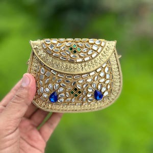 May include: A small, semi-circular gold-toned metal purse featuring an intricate embossed texture. The surface is embellished with numerous clear, reflective glass stones, two dark green square accents, and two prominent blue teardrop stones.