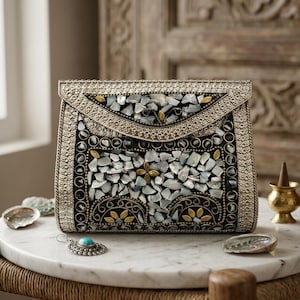 May include: A silver clutch bag with a black background, decorated with iridescent, irregularly shaped stones. The bag has detailed metalwork around the edges and a decorative flap. A turquoise earring and seashells are on the marble surface.