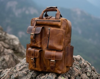 Könnte beinhalten: Ein hellbrauner Lederrucksack mit Tragegriff, einem Hauptfach mit Reißverschluss und vier Außentaschen mit Klappen, die durch Metallschnallen gesichert sind. Der Rucksack verfügt über zwei Seitentaschen und sichtbare Nähte am gesamten Design.