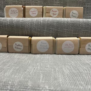 May include: An assortment of handmade soaps, each wrapped in brown paper and labeled with a white circular sticker. The labels feature names like "Nordic Night," "Highland Balsams," and "Tangier Jest." The soaps are arranged on a gray textured surface.