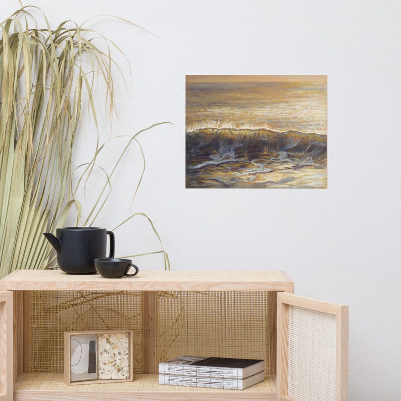 May include: An oil painting of ocean waves in shades of gold, brown, and blue. The artwork captures the movement of the water with a focus on the cresting wave. The painting is displayed on a white wall above a wooden cabinet.