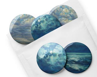 May include: A set of six round, glossy buttons with celestial-themed artwork. The buttons feature images of clouds, moons, and ocean waves in shades of blue, white, and gray. They are packaged in a white paper sleeve.