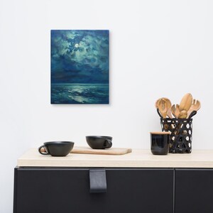 May include: An oil painting of a seascape at night, with a full moon and clouds. The painting is displayed on a white wall above a black cabinet. Two black mugs and a wooden cutting board are on the cabinet.