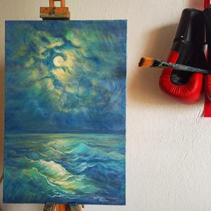 May include: An oil painting of a moonlit ocean scene in shades of blue and green, displayed on an easel. A pair of red and black boxing gloves hangs on the wall, with a paintbrush resting between them.