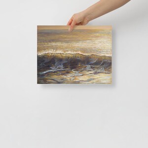 May include: A square print of an ocean wave, painted in shades of gold, brown, and blue. The wave crests with white foam, and the water reflects the sunlight. The artwork is held by a person.