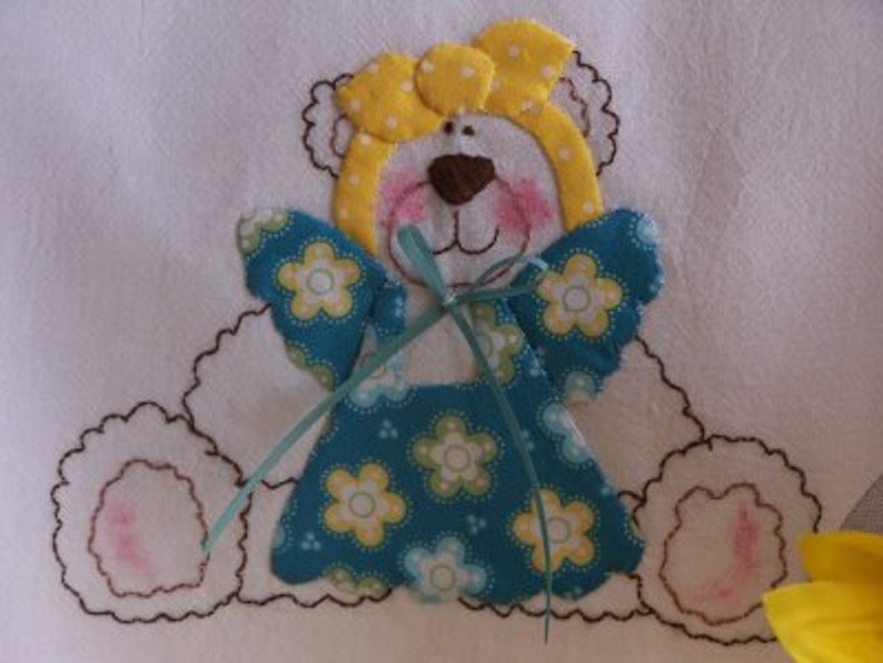 May include: A white fabric applique of a teddy bear with yellow polka dot bow, blue and white floral dress, and light blue wings.