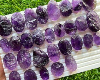 May include: A collection of polished amethyst gemstones in various shapes and sizes. The stones range in color from deep purple to light lavender, with visible crystalline structures. They are arranged on a white tray against a backdrop of green.