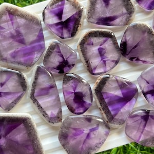 May include: A collection of polished amethyst stones with a range of shapes and sizes. The stones display a deep purple hue with white and black accents. The amethyst pieces are arranged on a white surface, showcasing their natural beauty.