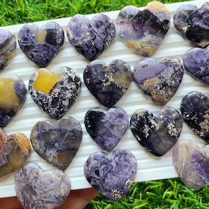 May include: A selection of heart-shaped stones in shades of purple, yellow, and brown. The stones are displayed on a white tray, highlighting their natural patterns and textures. The hearts vary in size and colour, creating a unique display.
