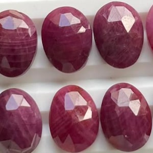 May include: A collection of oval-shaped, faceted ruby gemstones. The rubies display a deep red hue with visible internal patterns and a polished, reflective surface. A US quarter coin is included for size comparison.