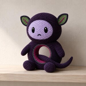 May include: A dark purple crocheted plush toy with a round head, a light purple face, and large black eyes. The body is circular with a hollow center, featuring a pink inner lining and a long, curved tail. The ears are green with pink accents.