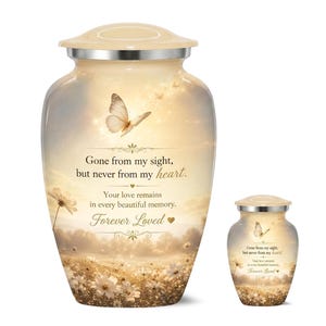 May include: A large and a small cremation urn featuring a cream and gold sunset landscape with a butterfly, white flowers, and the text: Gone from my sight, but never from my heart. Your love remains in every beautiful memory. Forever Loved.