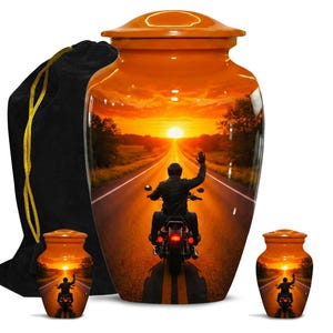 May include: Orange urn with a sunset scene of a person riding a motorcycle on a road. The urn has a matching small keepsake urn and a black velvet bag with a gold drawstring.