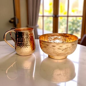 May include: A copper-colored Moscow Mule mug with a hammered texture and a handle, next to a gold-colored bowl with an ornate design. Both items are on a white surface, reflecting their shapes. The bowl has a decorative pattern around its rim.