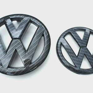 May include: Two carbon fiber Volkswagen emblems, one large and one small, are displayed against a white background. The emblems feature the iconic VW logo, with a textured, woven pattern. The larger emblem is approximately 6 inches in diameter.