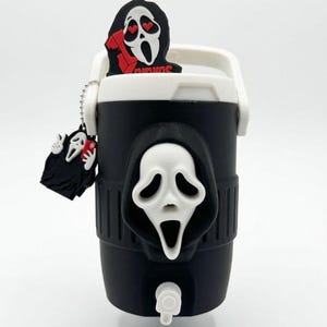 May include: A black and white beverage cooler featuring a prominent white ghost-like mask emblem on the front. The cooler includes a white handle and two decorative charms: one shaped like a mask with heart eyes and one showing a mask holding a phone.