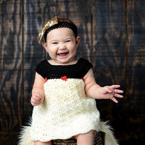 May include: A baby girl wearing a white crocheted dress with a black crocheted bodice and red bows. She is sitting on a brown textured stool with a white fur rug. She is wearing a gold glitter headband.