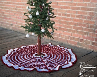 Puede incluir: Una falda de árbol de Navidad de crochet blanco y rojo con un borde festoneado. La falda está decorada con un patrón repetido de círculos rojos y blancos. Un pequeño árbol de Navidad artificial con adornos plateados se encuentra en el centro de la falda. Una media de Navidad de crochet blanco con rayas rojas cuelga de una pared de ladrillo en el fondo. El texto "Crafting Friends Designs" es visible en la esquina inferior derecha de la imagen.