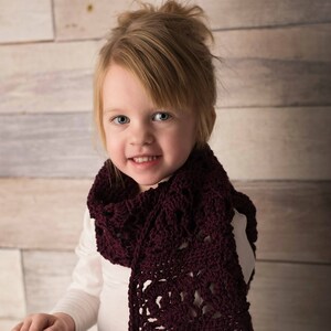 May include: A young girl wearing a white long-sleeved shirt and floral leggings is wearing a maroon crocheted scarf. The scarf has a lacy pattern and is tied around her neck.
