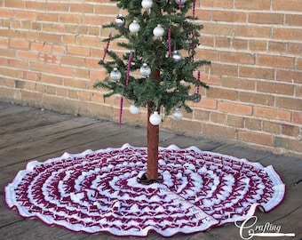 May include: A white and burgundy crochet tree skirt with a scalloped edge. The skirt is placed on a wooden floor in front of a brick wall. A small artificial Christmas tree with silver ornaments and a silver star on top is in the center of the skirt.