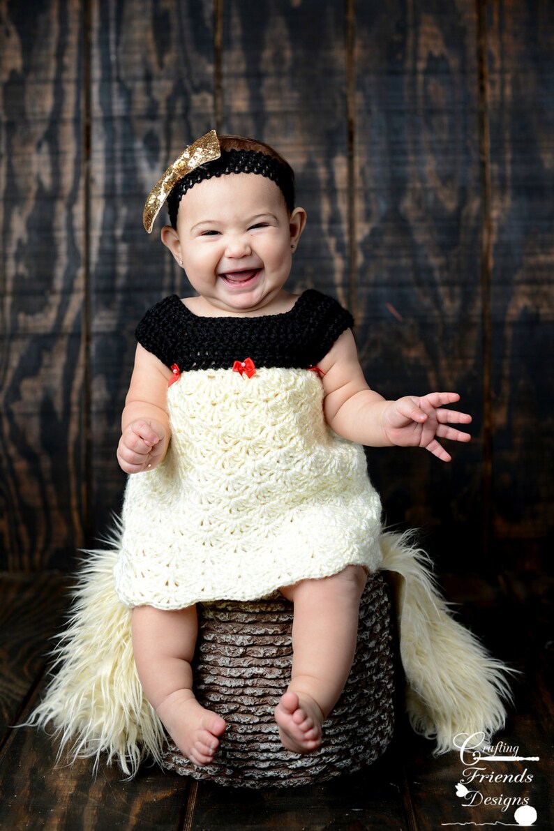 Peut inclure: Une petite fille portant une robe en crochet blanc avec un corsage en crochet noir et des n&oelig;uds rouges. Elle est assise sur un tabouret textur&eacute; brun avec un tapis en fourrure blanche. Elle porte un bandeau &agrave; paillettes dor&eacute;es.